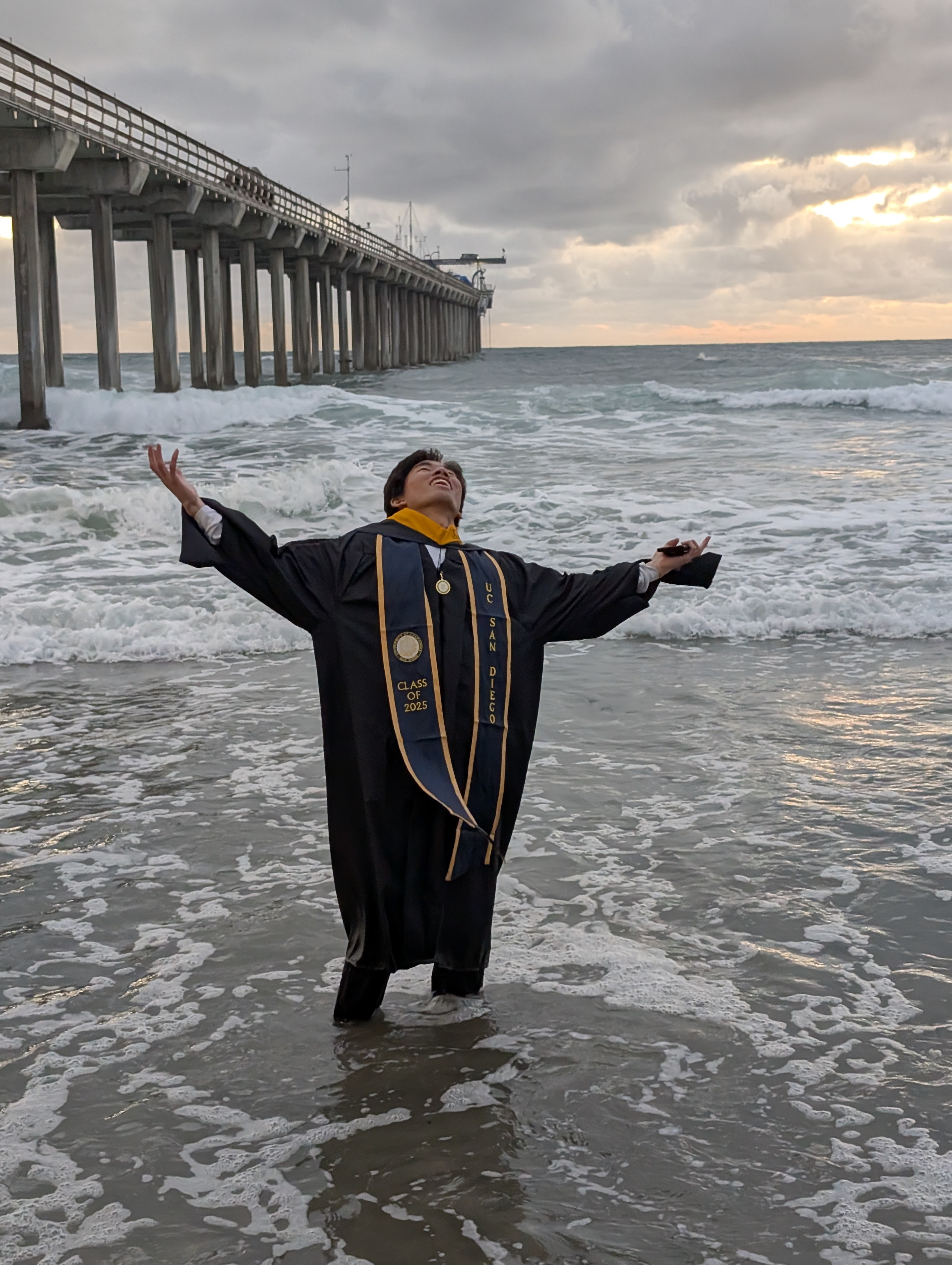 A photo taken by Alina of me triumphantly standing ankle deep in the Pacific Ocean, wearing a masters graduation robe, with Scripps Pier behind me, as the sun sets behind the clouds.